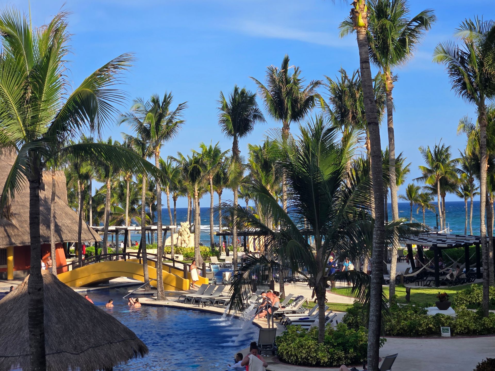 A resort scene with a swimming pool, yellow footbridge, thatched-roof structures, and palm trees overlooking the ocean.