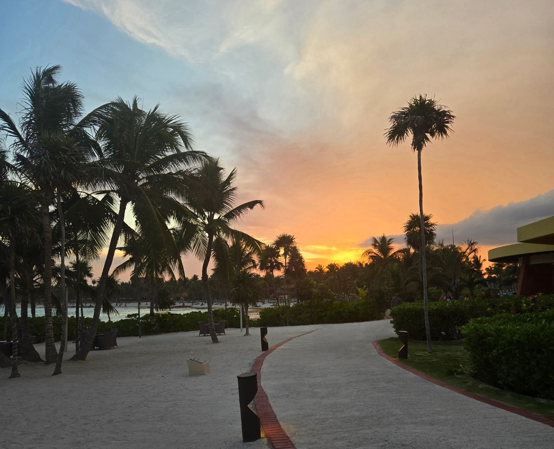 A white sandy path curves through palm trees toward a tropical beach under a vibrant sunset with orange and blue skies.