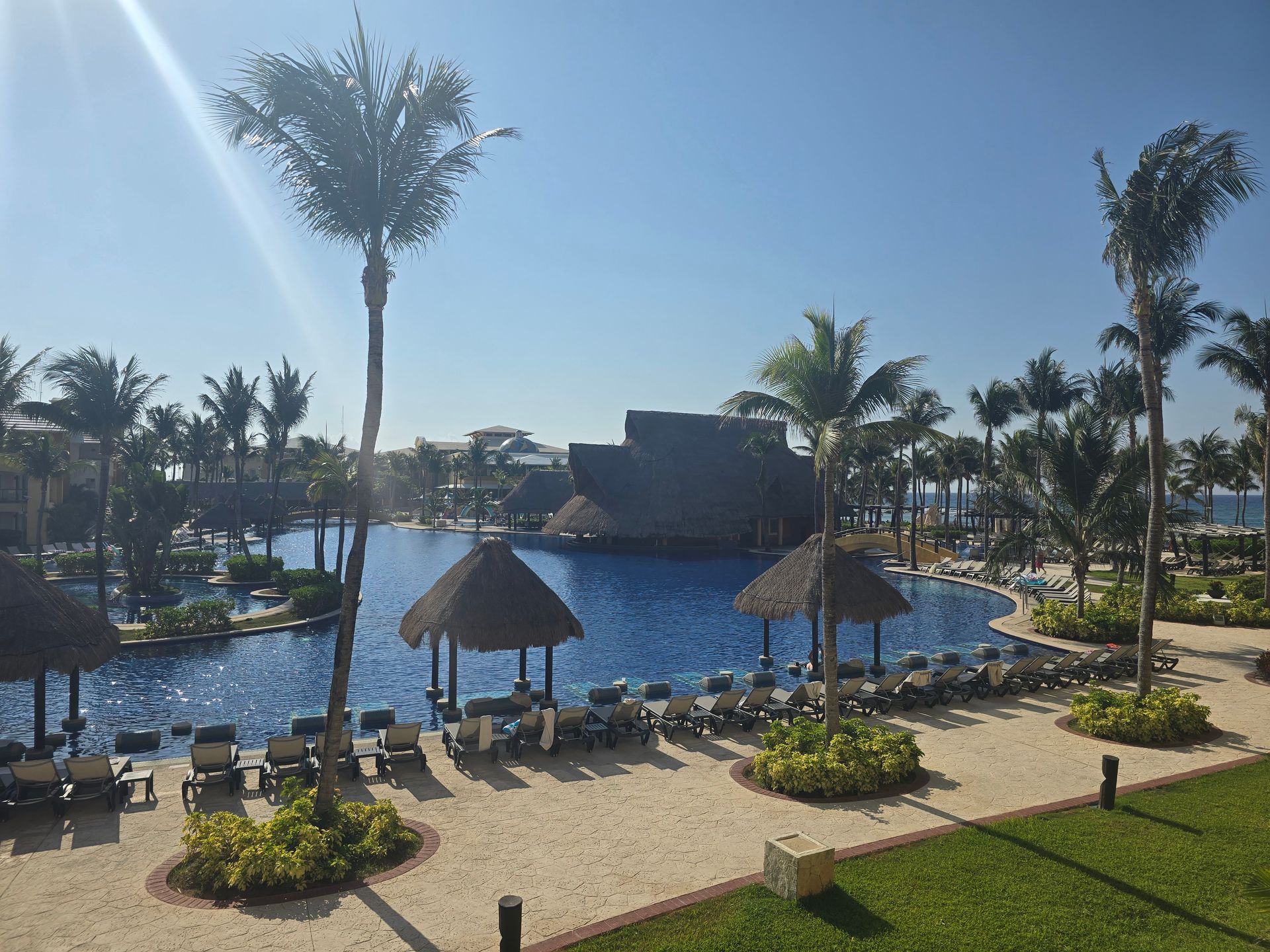 A sunny resort pool area with palm trees, thatched-roof cabanas, and lounge chairs on a sandy patio overlooking the sea.