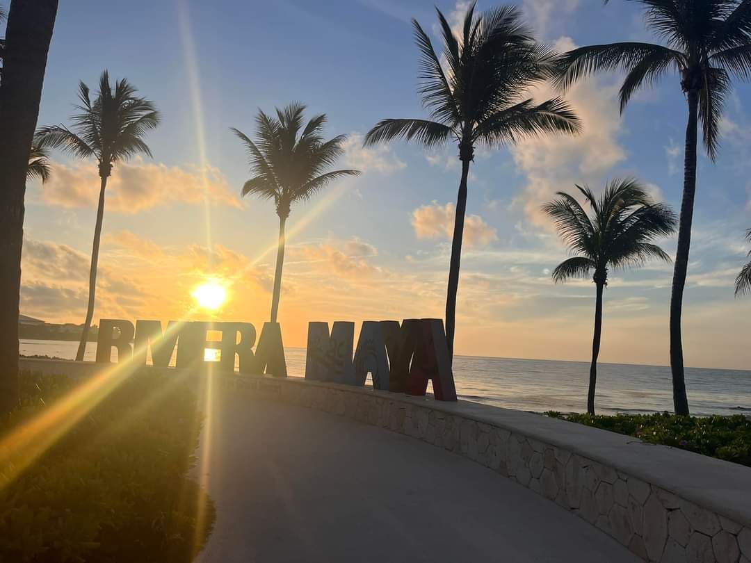 A sunset over the ocean in Riviera Maya, with palm trees silhouetted along a paved walkway beside a large name sign.