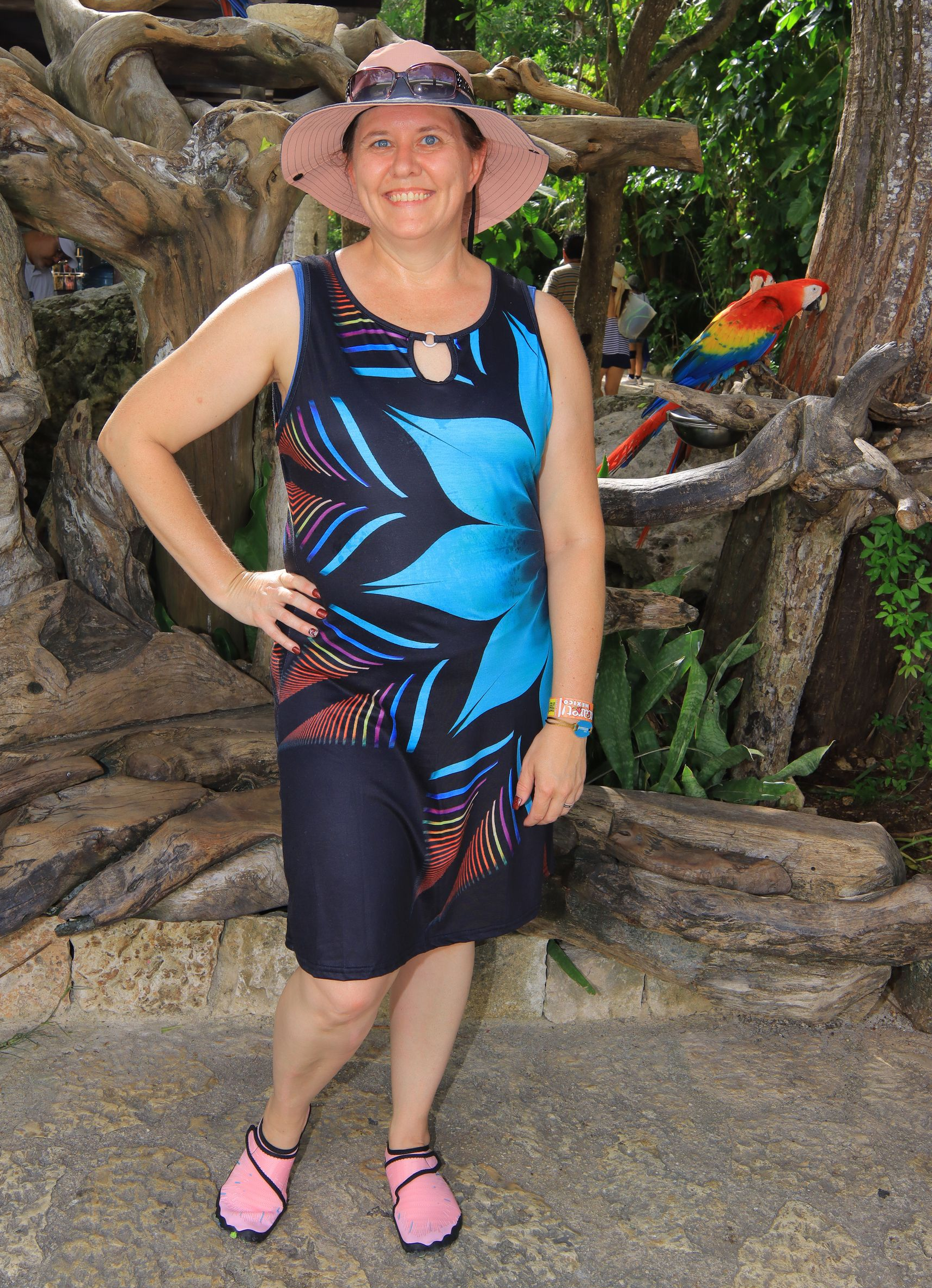 A smiling person in a blue and black patterned dress and pink hat stands in a lush outdoor setting with a colorful macaw.