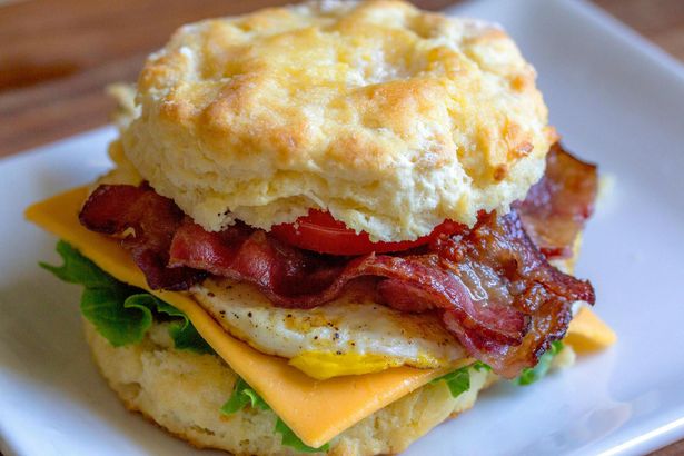 A biscuit sandwich with bacon , cheese , eggs and lettuce on a white plate.