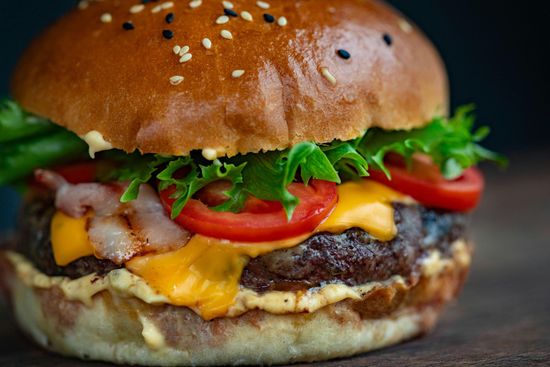 A close up of a hamburger with cheese , lettuce , tomatoes and bacon on a wooden table.