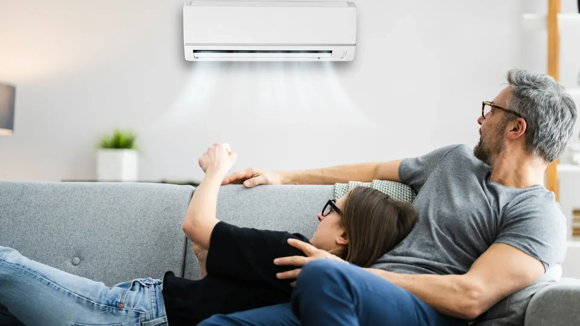A man and a woman are laying on a couch under an air conditioner.