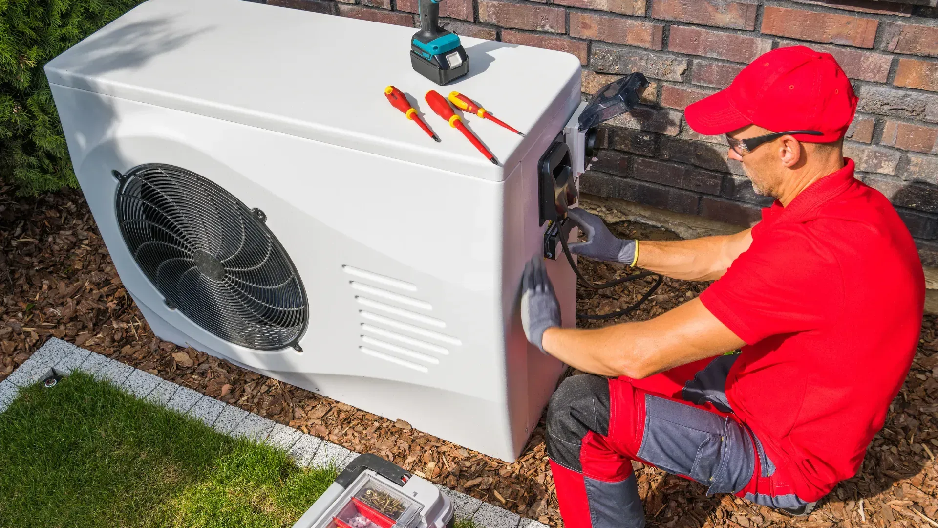 A man is sitting on the ground working on a heat pump.