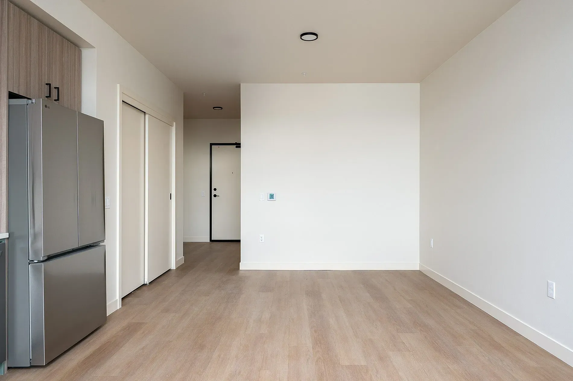 Empty apartment living room and kitchen area with stainless steel refrigerator and closet doors.