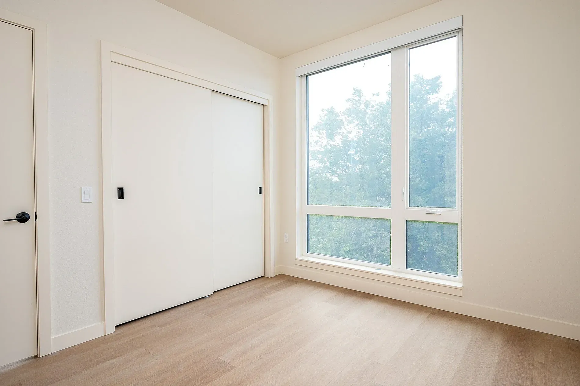 Empty bedroom with sliding closet door and large window with trees outside.