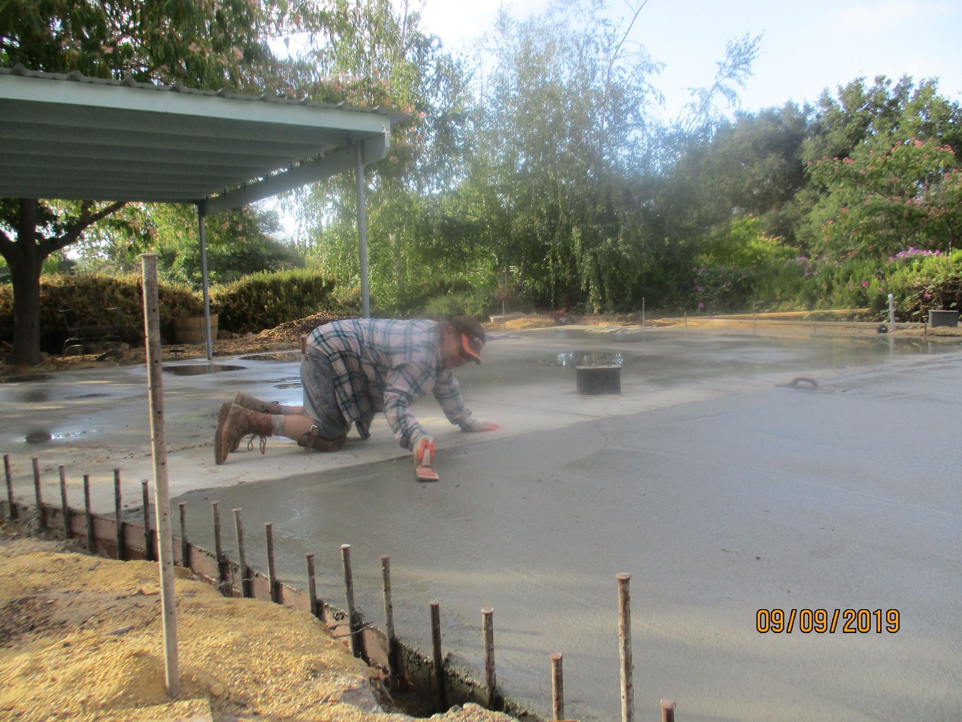 Person kneeling on a wet driveway, spraying water from a hose near a carport with trees in the background