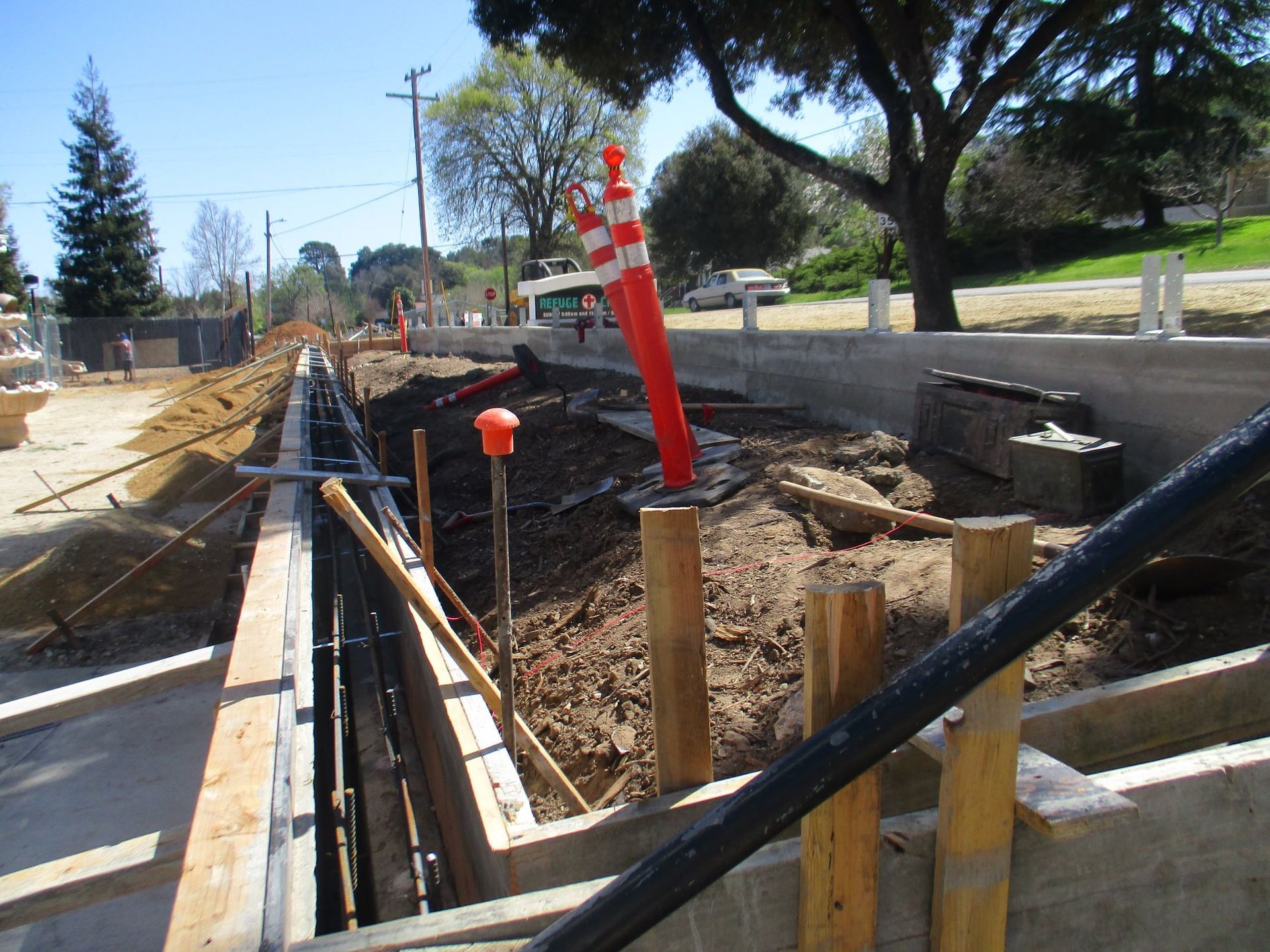 Road construction trench with orange safety markers, wooden supports, and workers’ equipment beside a street.