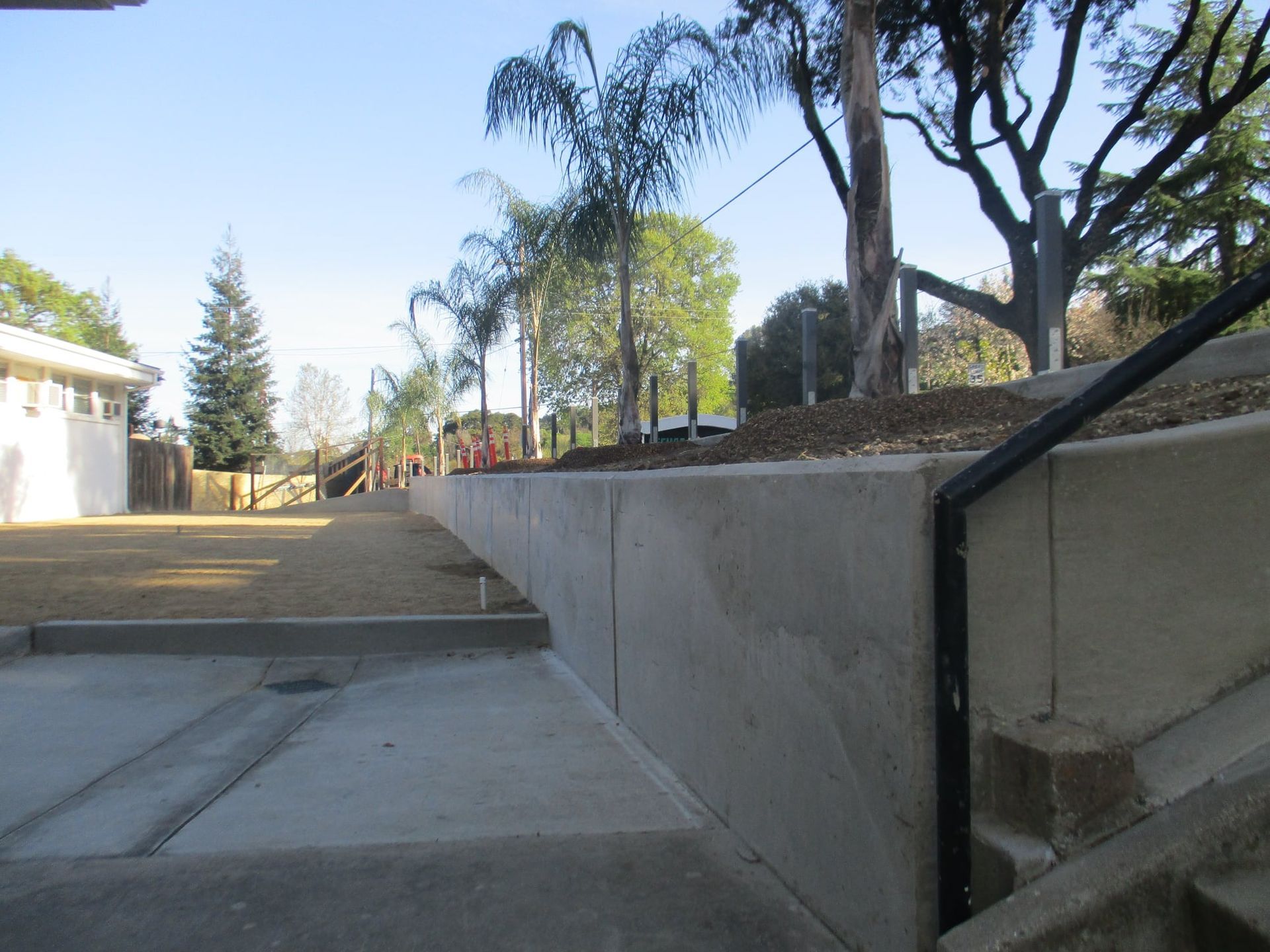 Concrete walkway and stairs beside a low wall with palm trees and a house in the background