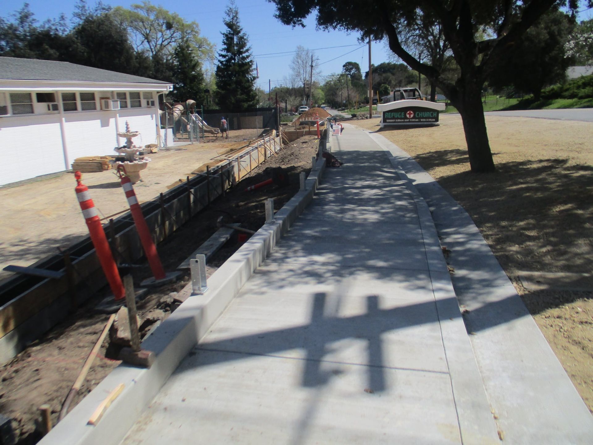 Sidewalk beside a construction trench, with orange barricades and a tree-lined street nearby