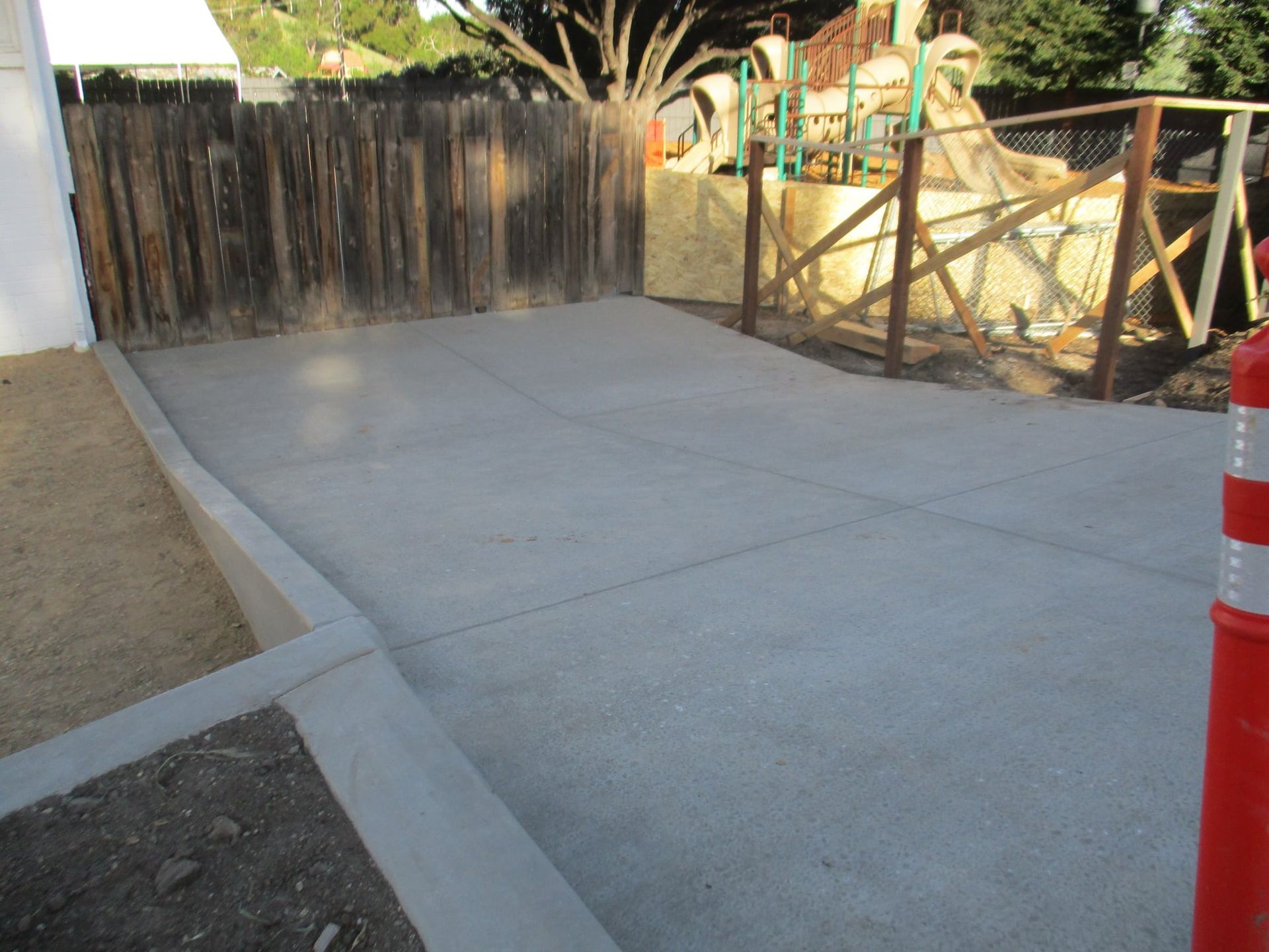 Concrete driveway beside a wooden fence and gate, with an orange traffic cone at right.