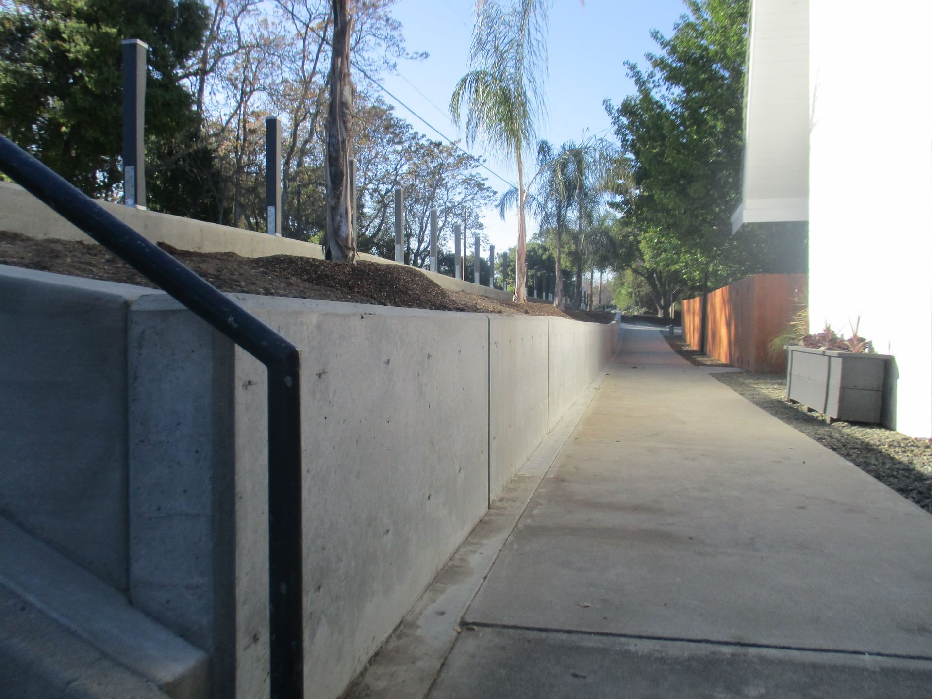 Concrete walkway beside a retaining wall and stairs, leading past trees and a building in daylight