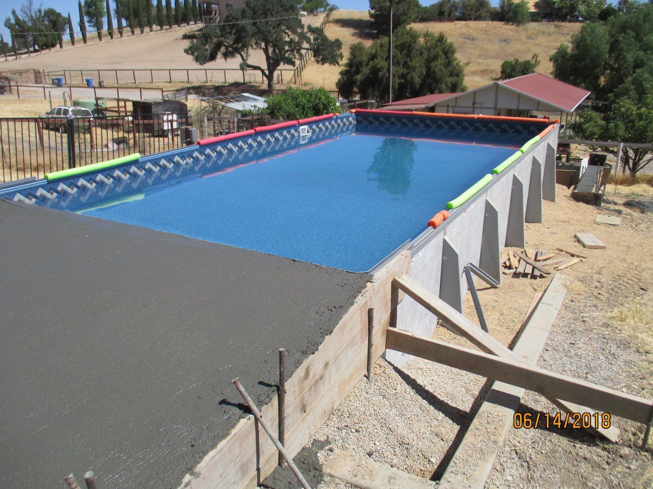 Outdoor swimming pool under construction beside a dirt path and rural hillside, with blue water and red edging