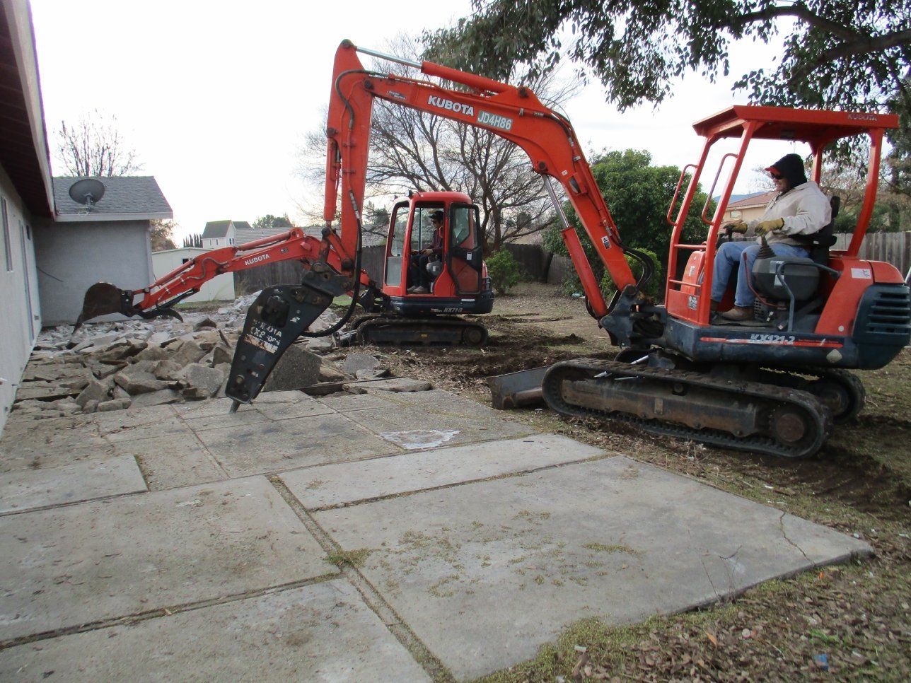 Two red excavators digging concrete in a yard, with one operator seated in the right machine.