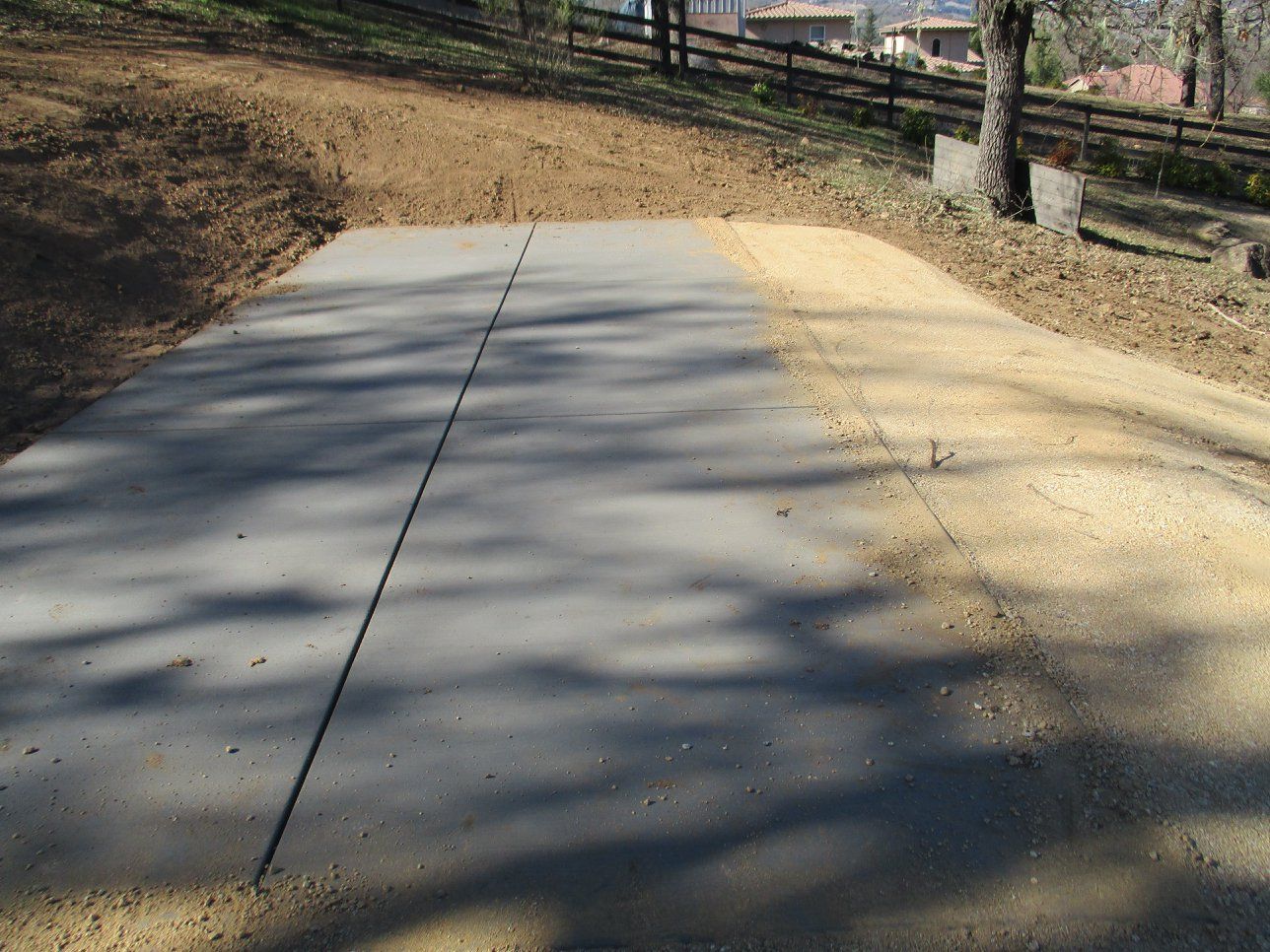 Curved concrete sidewalk beside a tan gravel path with trees and fence shadows