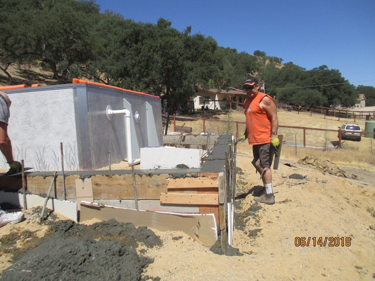Construction worker in orange vest stands beside foundation walls at a building site.