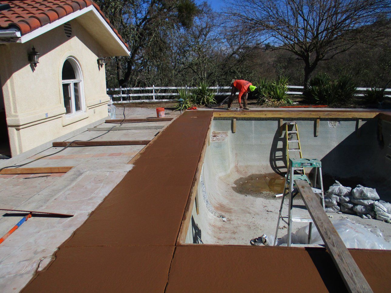 Worker resurfacing a pool deck beside an empty swimming pool and house