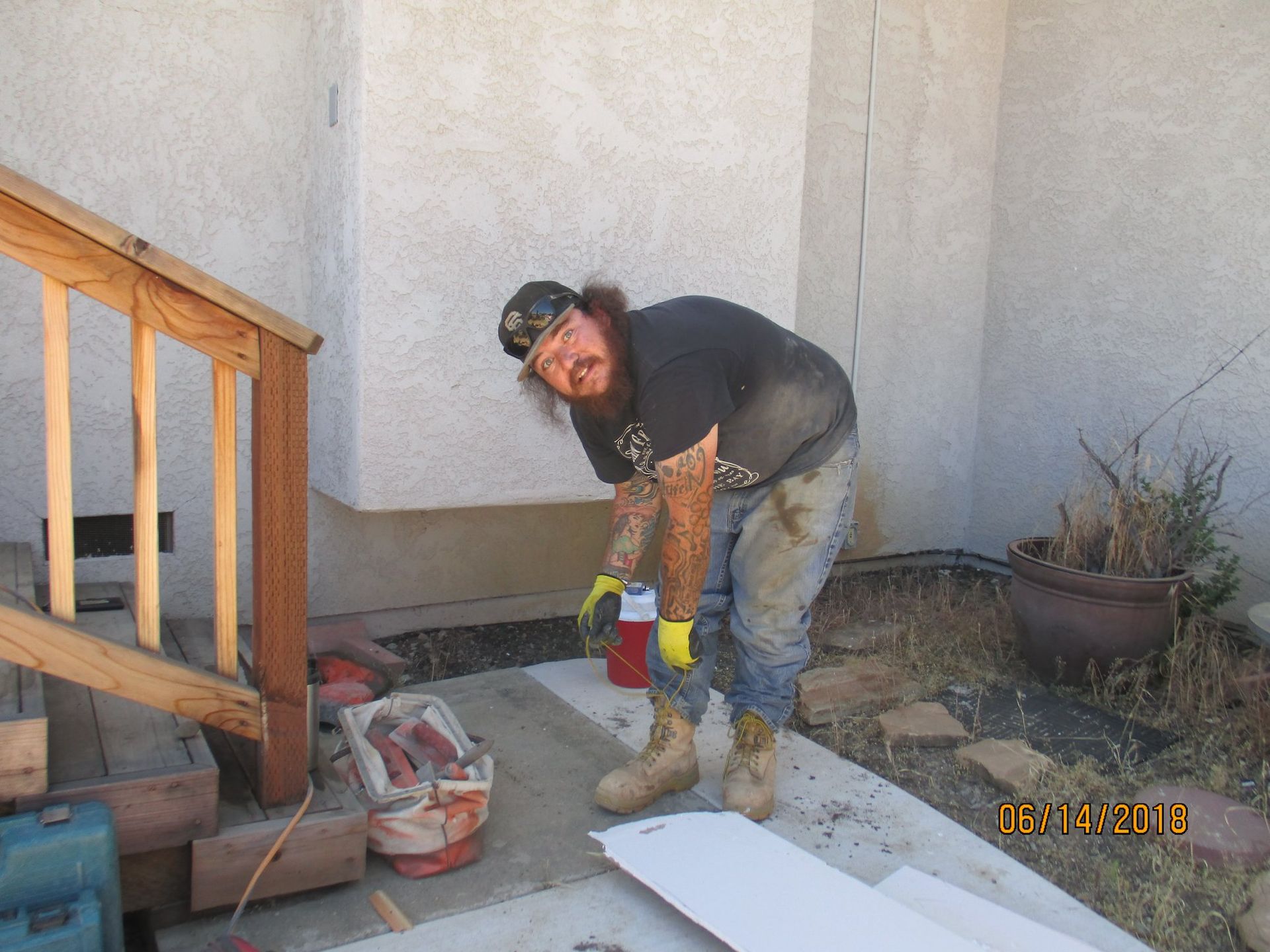 Worker in black shirt and jeans shoveling concrete mix on a porch next to stairs and a wall.