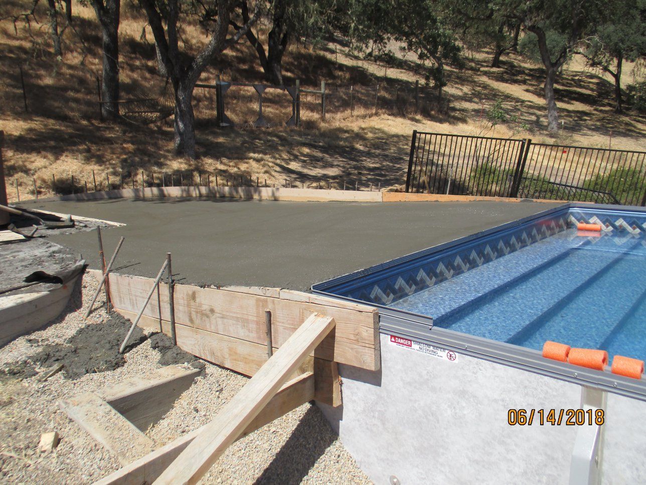 Concrete pool deck beside blue pool under construction, with wooden formwork and gravel road nearby.