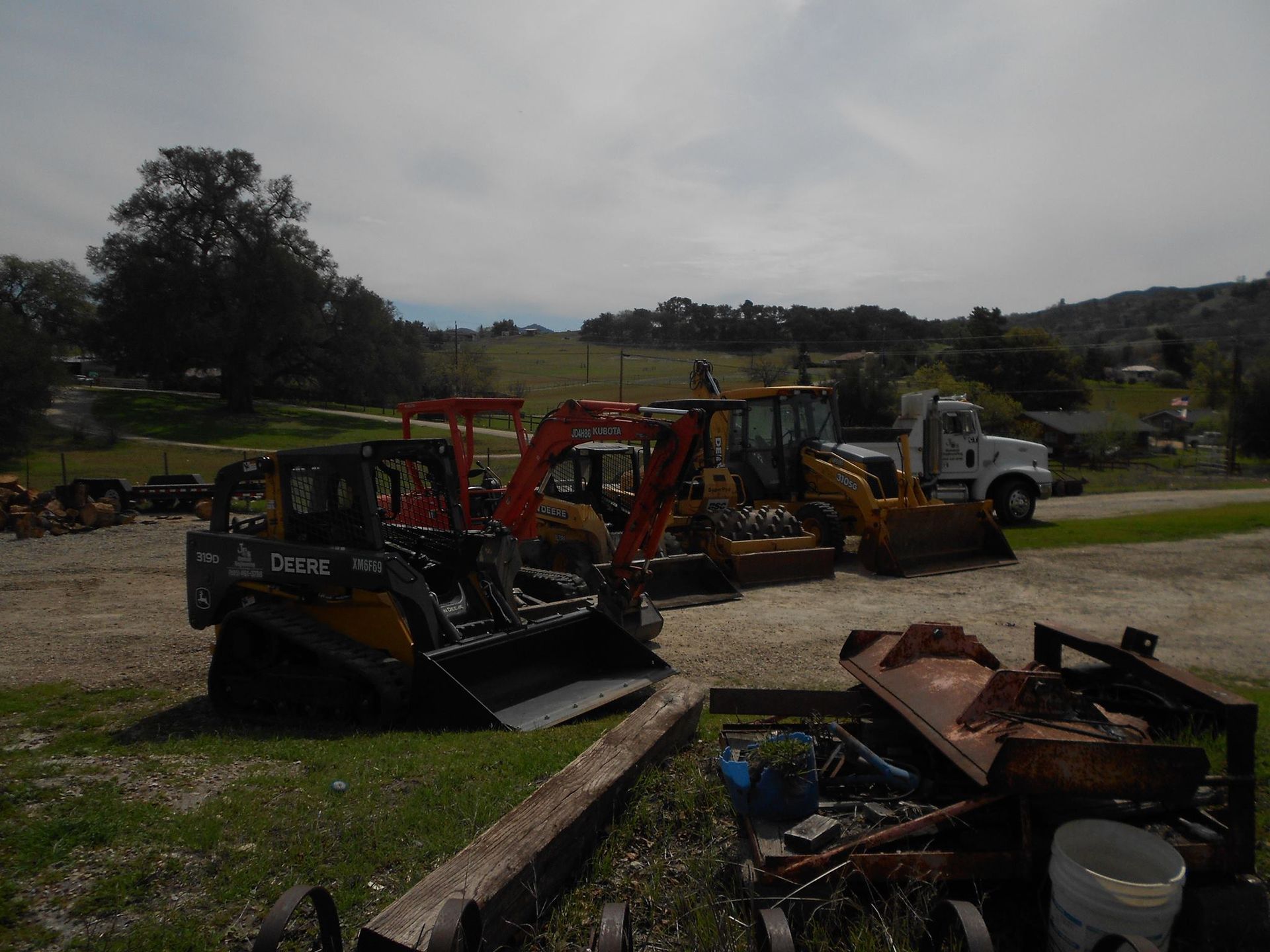 Construction equipment parked on a gravel lot beside a grassy field under a cloudy sky