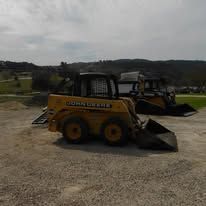 Yellow John Deere backhoe loader parked on a gravel lot with a bucket attachment and hills in the background