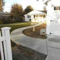 Curved concrete walkway through a yard leading to a white house and gazebo in a fenced backyard.