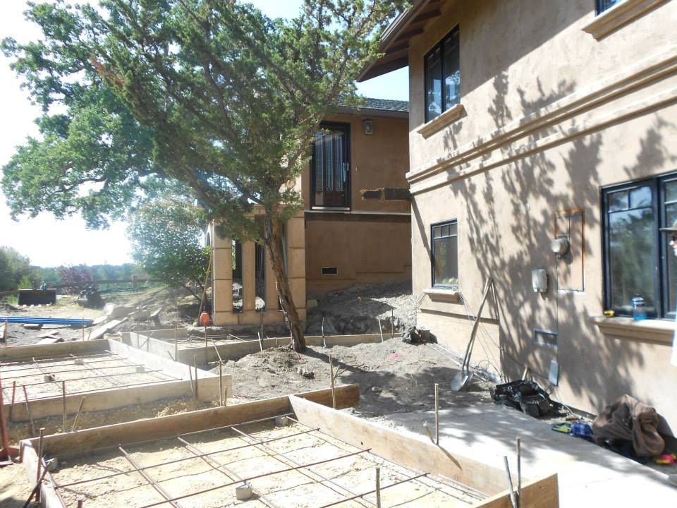 Outdoor construction site beside a beige building with wooden forms, concrete slabs, and a large tree casting shade