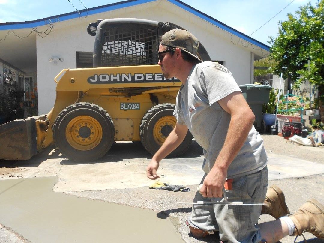 Man in gray shirt crouches near a yellow John Deere loader in a sunny driveway.