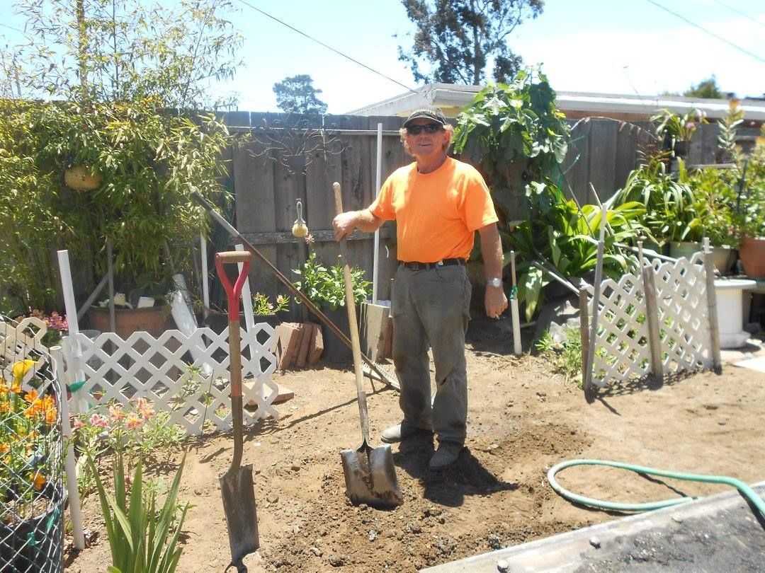 Person in orange shirt stands in a garden holding a shovel beside a hose and white trellis fence.