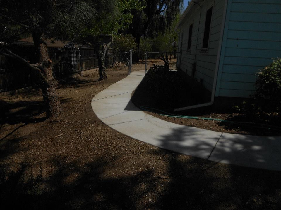 Curved concrete walkway beside a house, bordered by dirt and trees in a shaded yard.