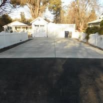 Wide driveway and white garage with paved parking area, flanked by white fences and trees.