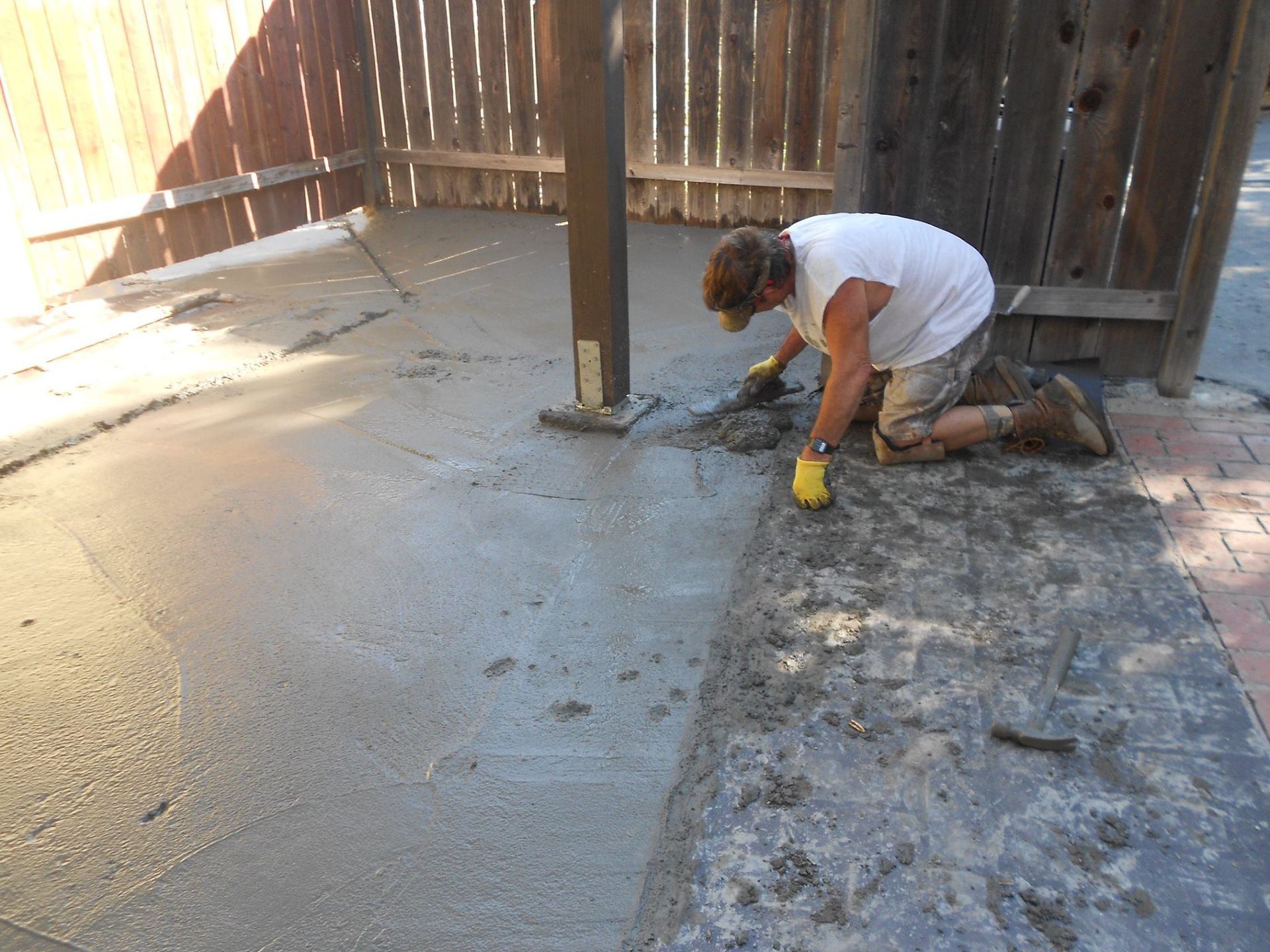 Worker troweling wet concrete beside a wooden fence and post in a backyard.