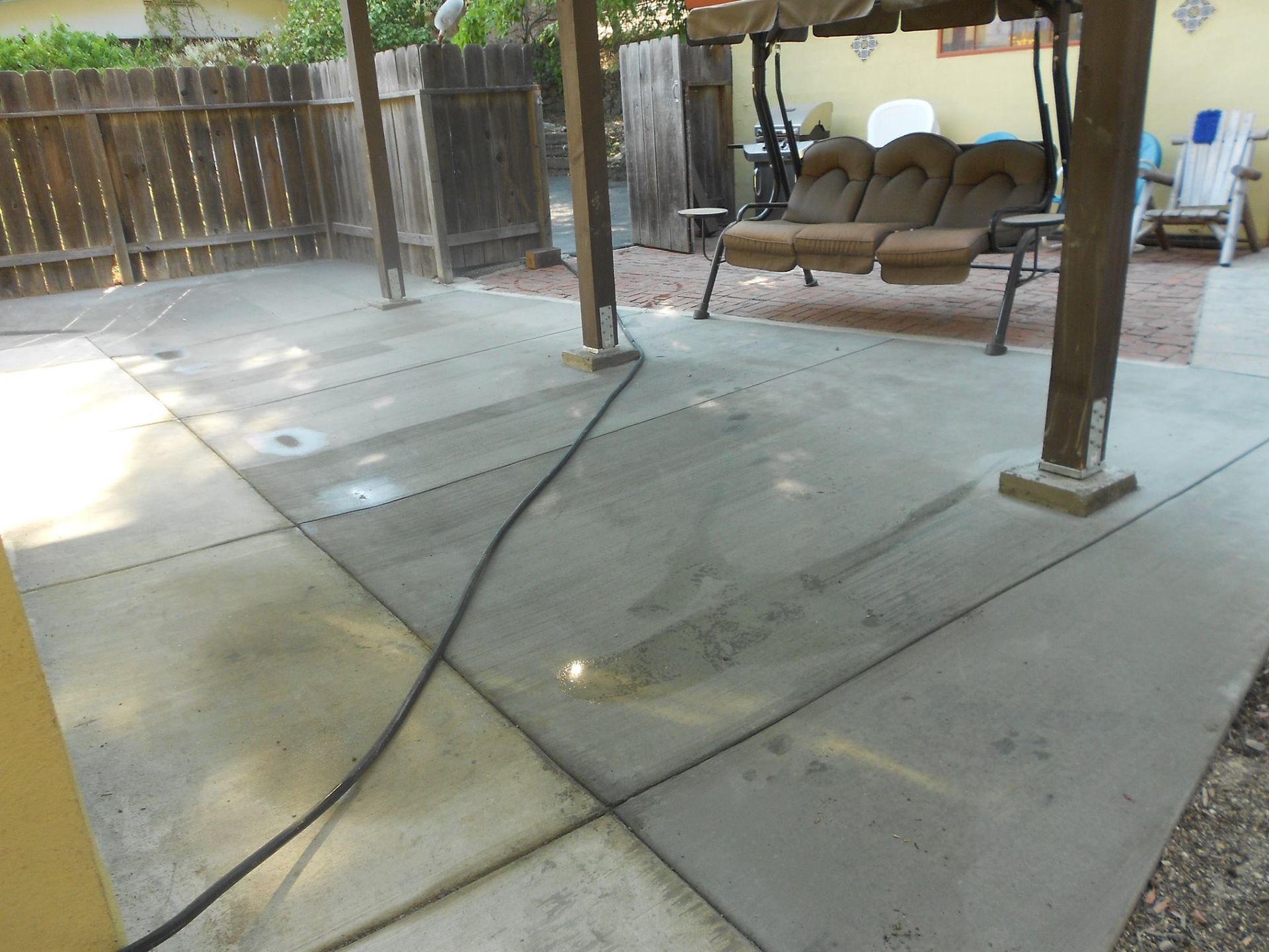 Covered backyard patio with concrete floor, swing bench, wooden posts, and a hose on the ground.
