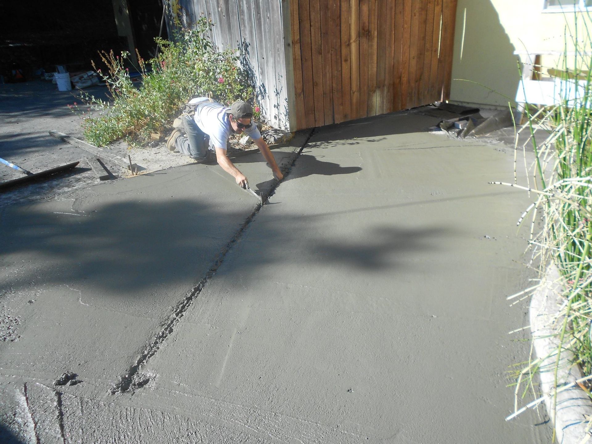 Person crouching on a concrete driveway near a wooden fence, working on a crack with a tool.