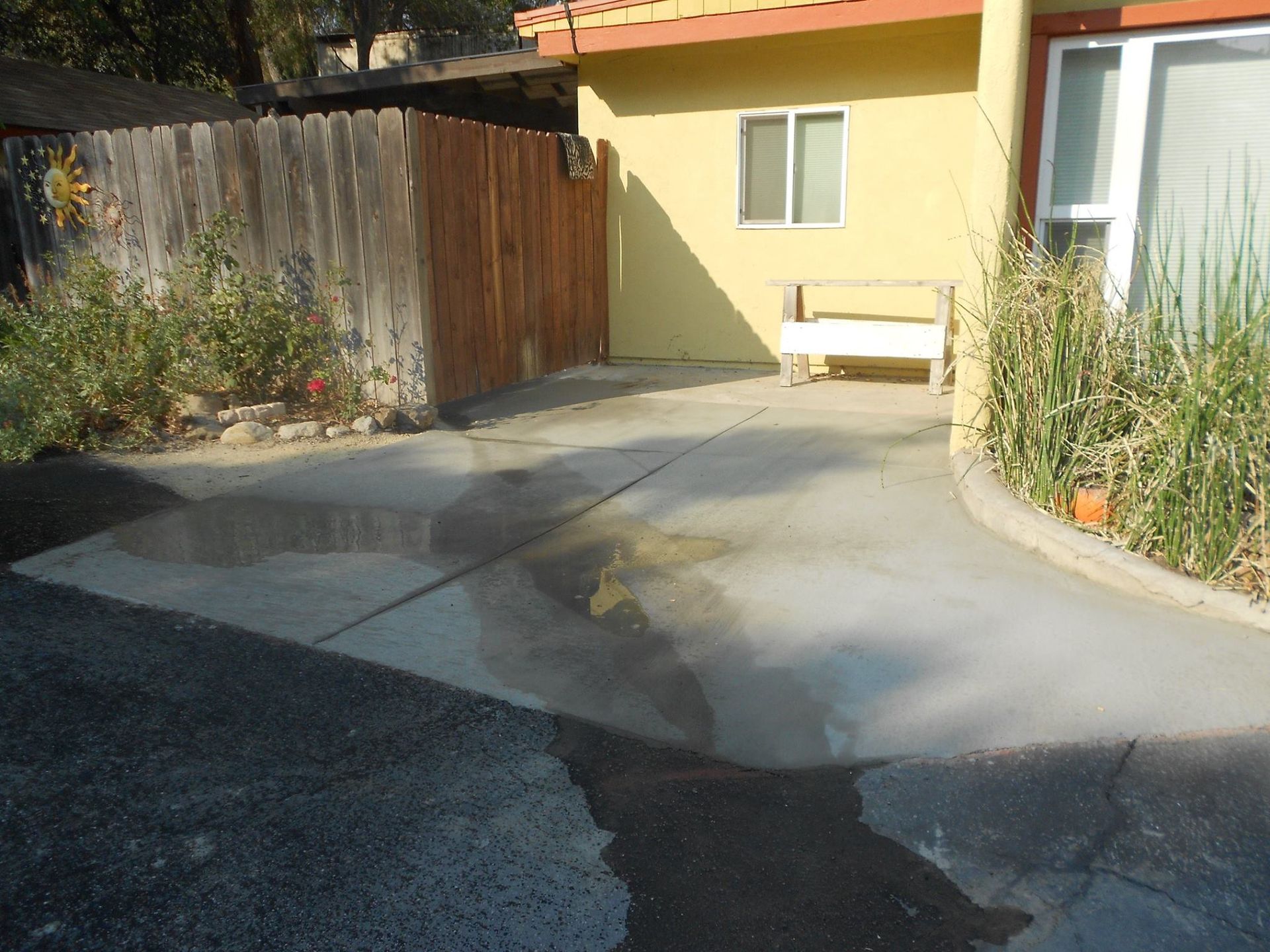 Sunny driveway beside a yellow house, with a wooden fence, bench, and tall plants along the wall.