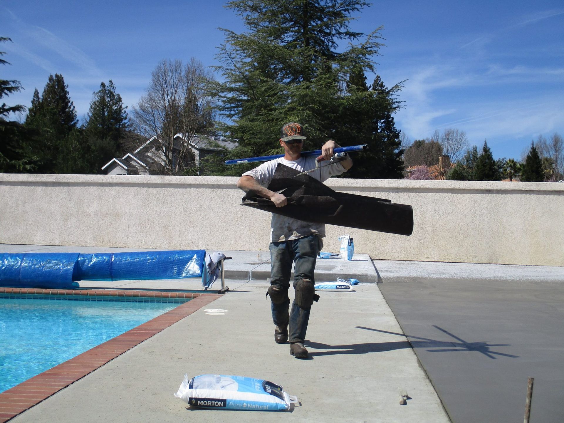 Person standing by a pool holding a long black object, with trees and houses in the background.