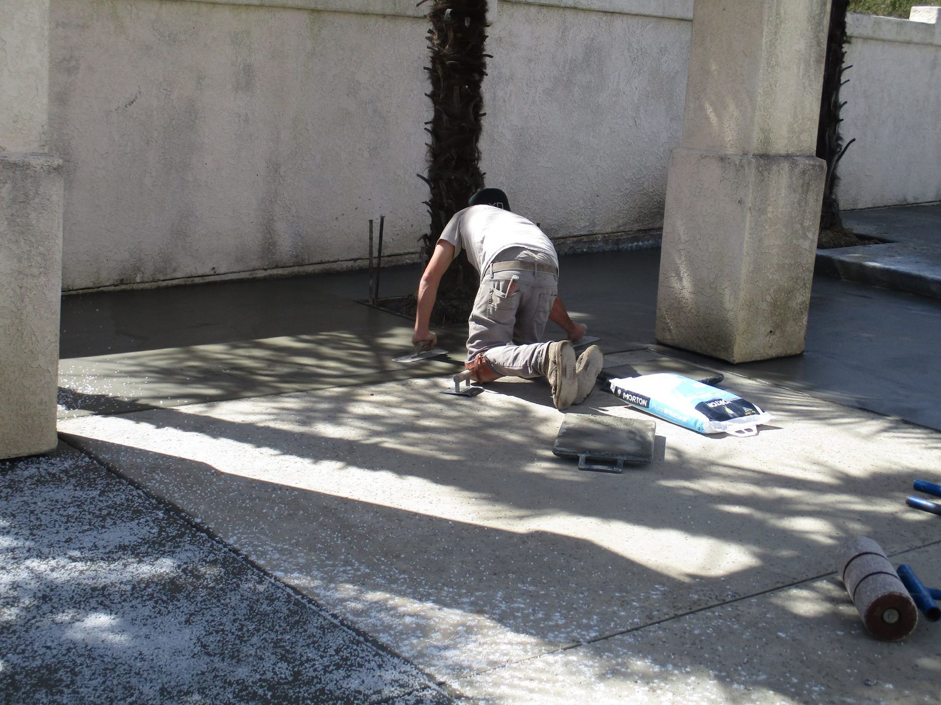 Worker kneeling on a concrete patio, handling tools beside a wall in bright sunlight
