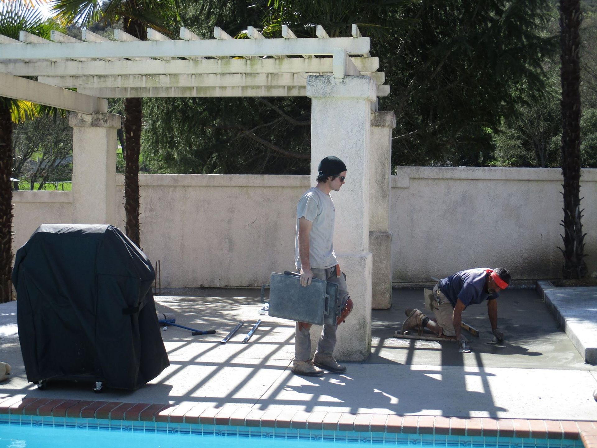 Two people work in a sunlit patio beside a white pergola and wall; one stands, one kneels near tools.