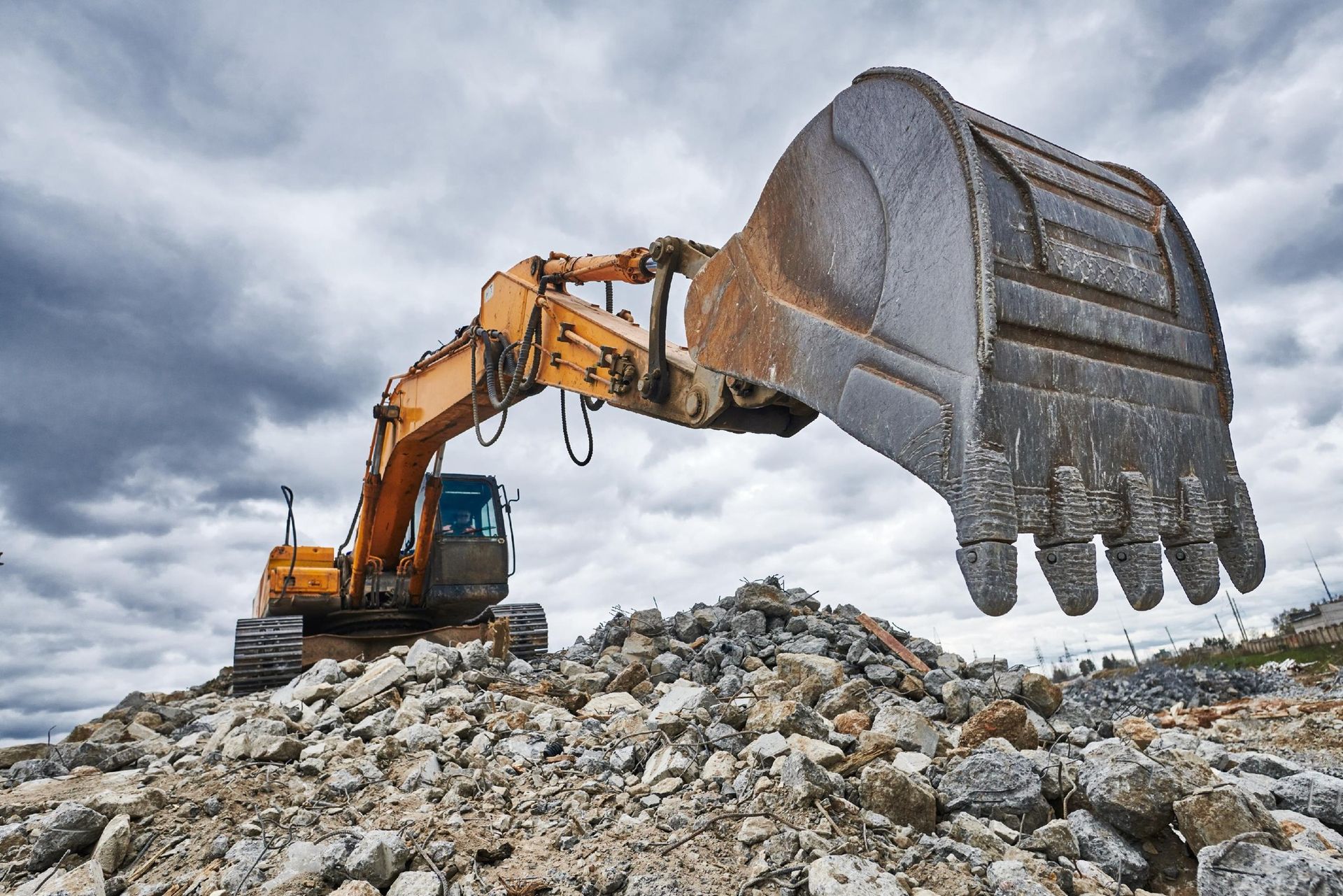 Orange excavator with raised bucket on a rocky construction site under cloudy sky