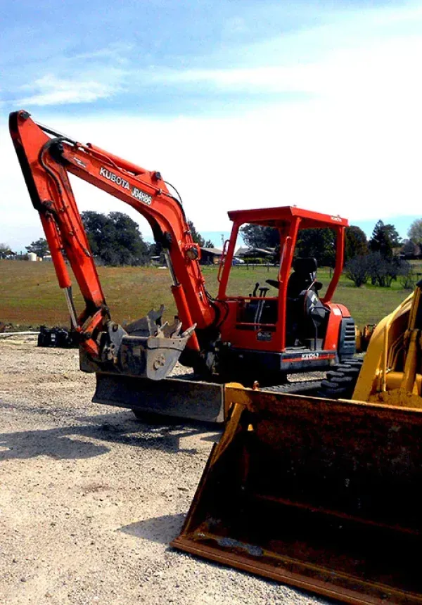 Orange mini excavator and yellow loader bucket parked on a dirt lot under a clear sky
