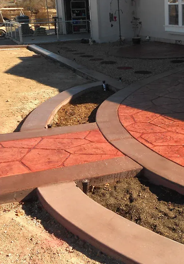 Curved red concrete walkway and steps in a landscaped yard beside a house.