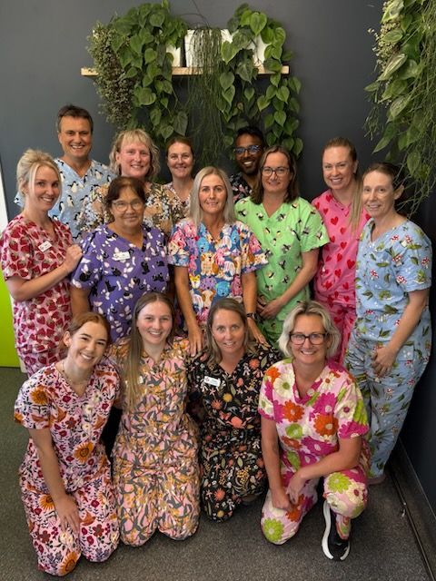 A group of 14 people in patterned scrub tops smiling in front of a dark wall with hanging plants. — Kanwal Veterinary Hospital In Kanwal, NSW