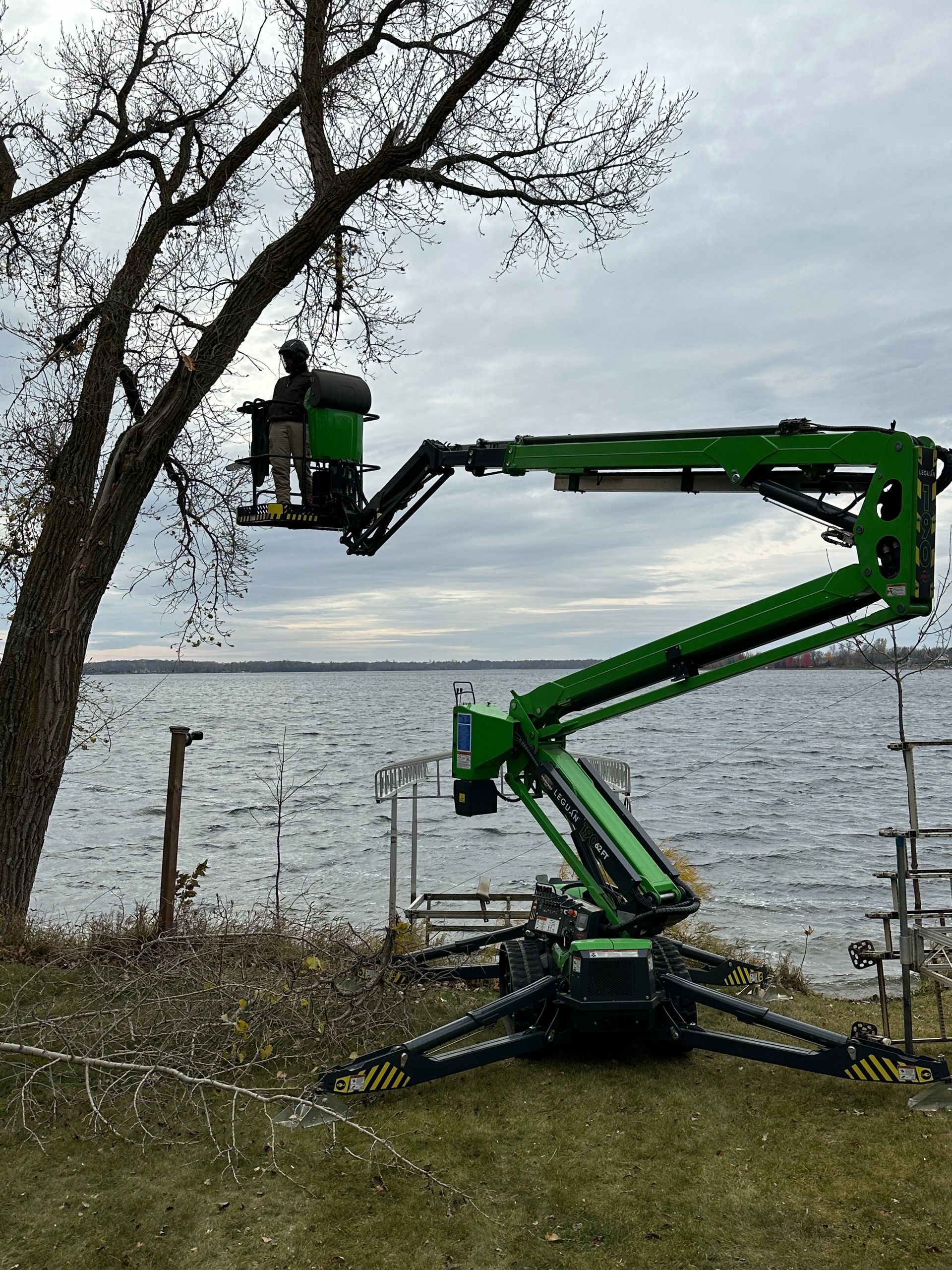 A green boom lift with a worker trimming a tree next to a lake. Overcast sky. A green boom lift with a worker trimming a tree next to a lake. Overcast sky.