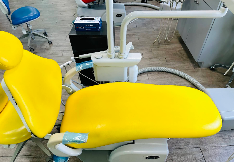Yellow dental chair in a clinic. Blue and white dental tools and chair, gray cabinets, and blue chair in background.