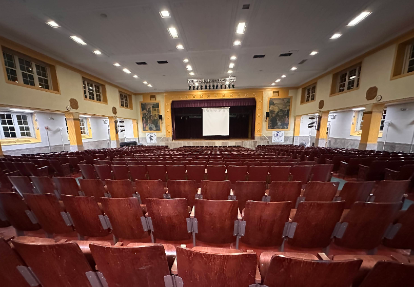 An empty auditorium with rows of brown seats facing a stage with a screen.