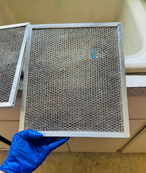A person holding a greasy, metal range hood filter. Blue glove. Kitchen setting.