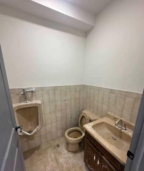 Small, outdated bathroom with a urinal, toilet, and sink; off-white walls and tile, poor condition.