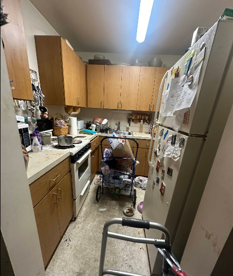 Small, cluttered kitchen with an old refrigerator covered in papers. A walker is in the foreground.