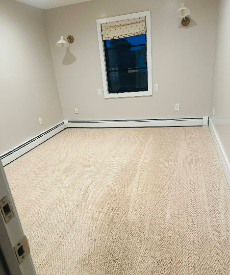 Empty bedroom with beige carpet, tan walls, window with curtain, and wall sconces.